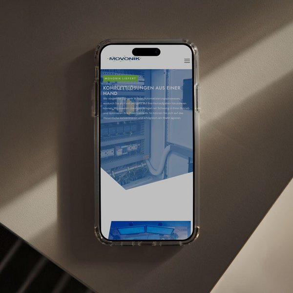 Smartphone im Hochformat auf einer glatten Oberfläche zeigt Movonik-Webseite mit dem Slogan „KOMPLETTLÖSUNGEN AUS EINER HAND“.