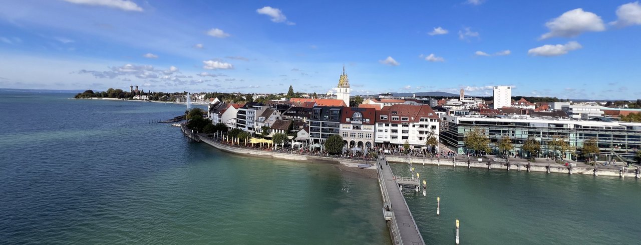 Uferpromenade einer Bodenseestadt: rote Dächer, goldene Turmspitze, Café-Terrassen und Fußgängerbrücke ins Zentrum.