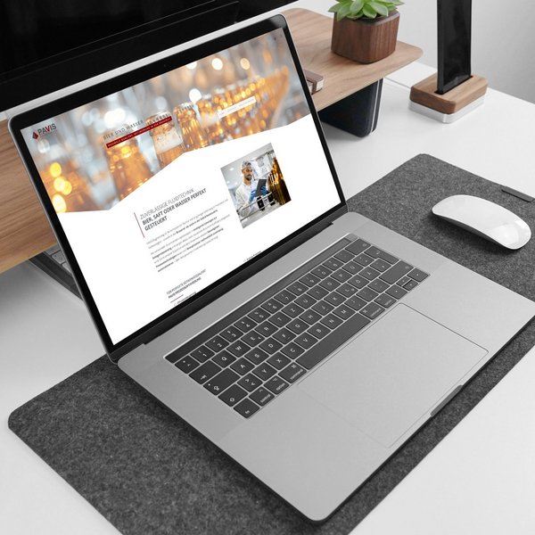 Laptop am Schreibtisch, Maus, Uhr und Pflanze daneben; Bildschirm zeigt eine Bier-Website mit Gläsern.