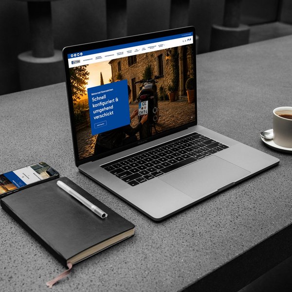 Laptop zeigt eine Website, daneben Smartphone, Notizbuch mit Stift und eine Tasse Kaffee auf grauem Tisch.