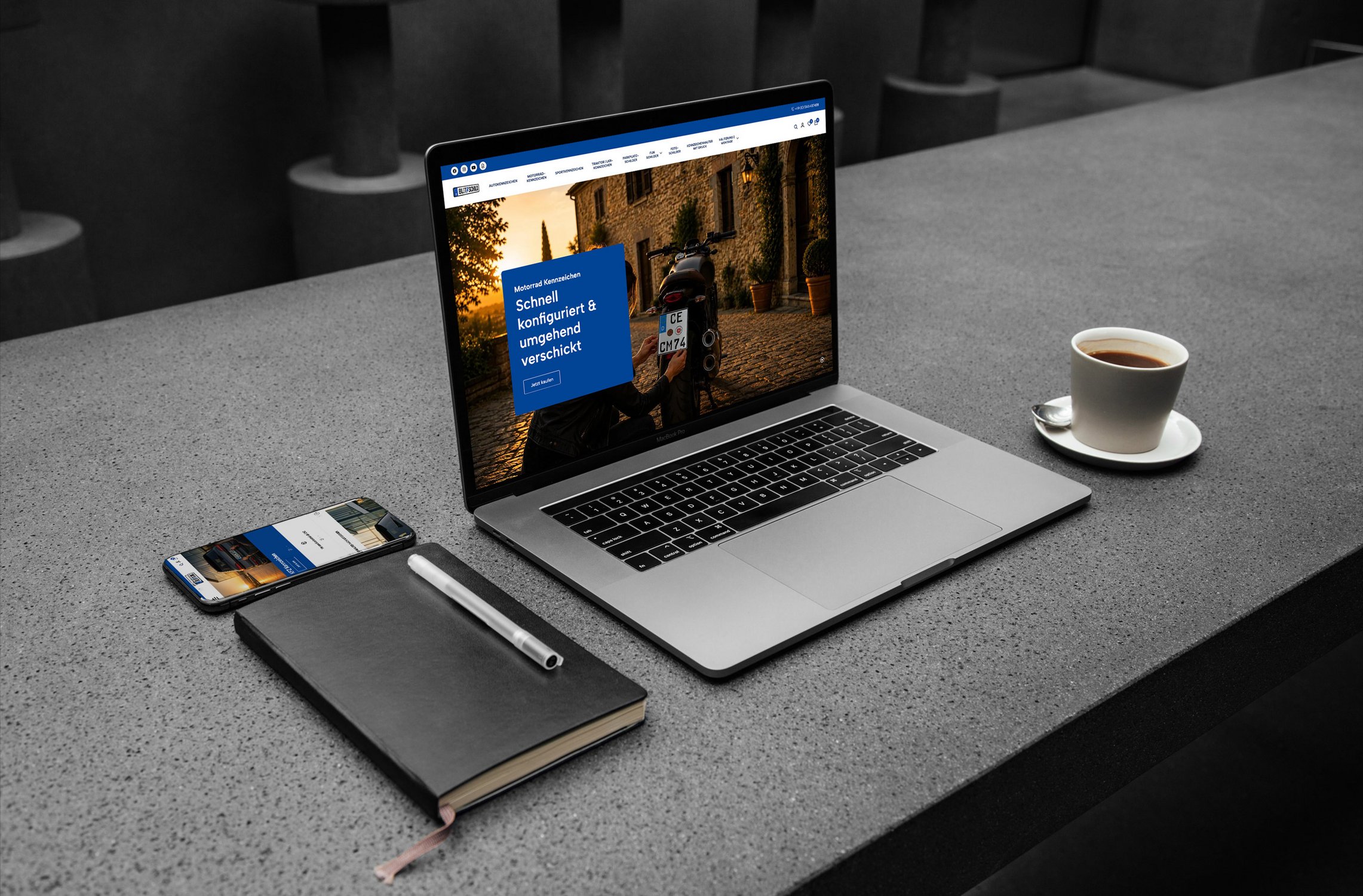 Laptop zeigt eine Website, daneben Smartphone, Notizbuch mit Stift und eine Tasse Kaffee auf grauem Tisch.
